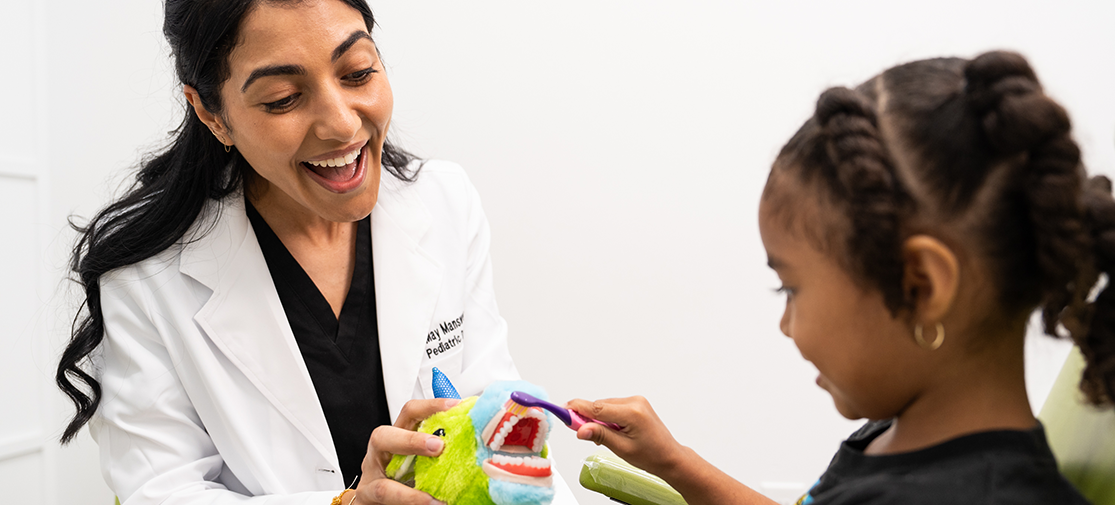 Dr. May Manswer showing a patient how to brush her teeth using stuffed animals as an example